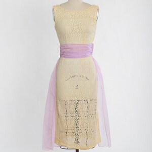 Op de afbeelding: Een gele kanten jurk met een lavendelkleurige band en een transparante lavendelkleurige overlay.