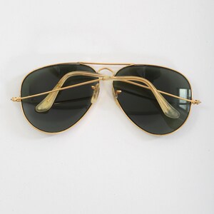 May include: Aviator sunglasses featuring a thin gold-tone metal frame with a double bridge design. The lenses are dark green, and the temples are finished with translucent, light-colored plastic ear pieces that curve inward toward the center.
