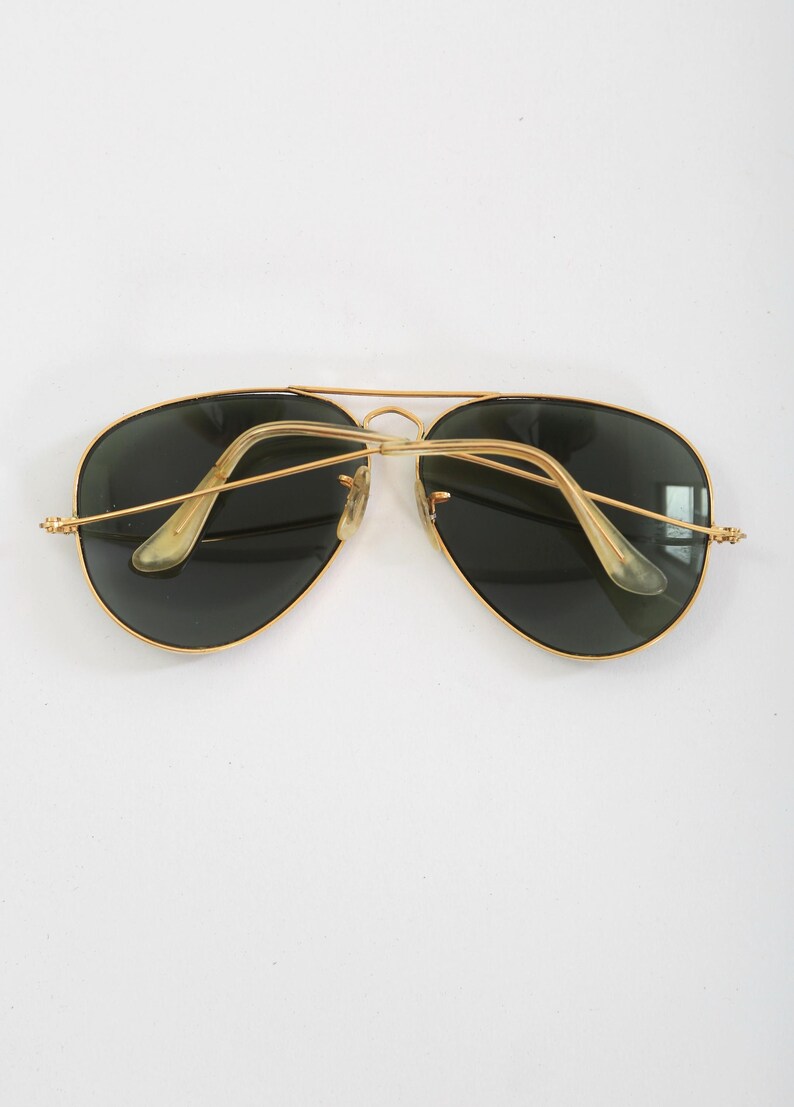 May include: Aviator sunglasses featuring a thin gold-tone metal frame with a double bridge design. The lenses are dark green, and the temples are finished with translucent, light-colored plastic ear pieces that curve inward toward the center.