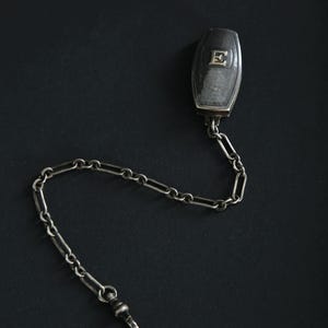 May include: A silver-toned pocket watch chain with a rectangular fob featuring the letter "E". The chain is composed of small, interconnected links and a clasp. The fob and chain are set against a dark background.
