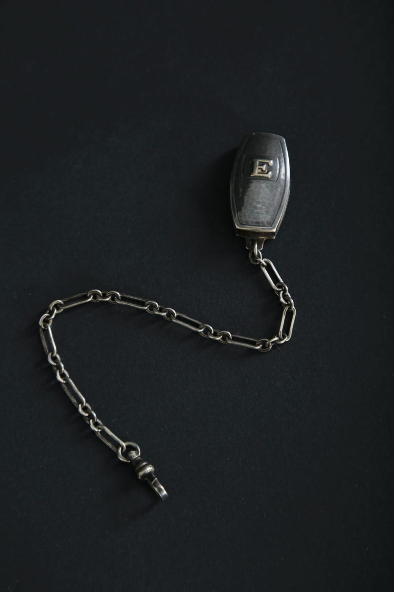 May include: A silver-toned pocket watch chain with a rectangular fob featuring the letter "E". The chain is composed of small, interconnected links and a clasp. The fob and chain are set against a dark background.