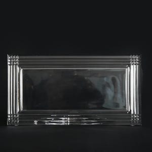 May include: A rectangular, clear glass tray with a decorative, ridged border. The tray has a simple, elegant design, suitable for serving or display. The tray is approximately 10 inches long and 6 inches wide.
