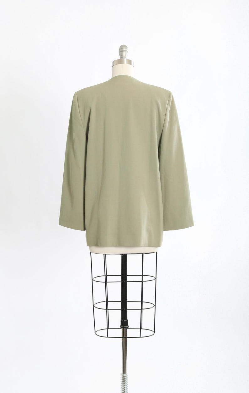K&ouml;nnte beinhalten: Ein hellgr&uuml;ner Blazer mit langen &Auml;rmeln, auf einer Schneiderpuppe pr&auml;sentiert. Der Blazer hat ein schlichtes, klassisches Design und eine glatte Textur. Die Schneiderpuppe ist schwarz-wei&szlig;, mit einem Drahtk&auml;fig-Design.