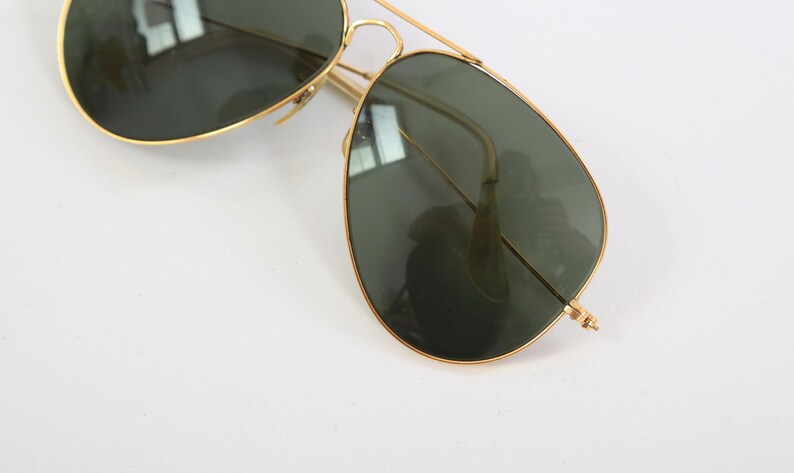 May include: Aviator sunglasses featuring a thin gold-tone metal frame and dark green teardrop-shaped lenses. The design includes a double bridge across the top and slender temples, providing a classic look with a metallic finish and tinted glass.