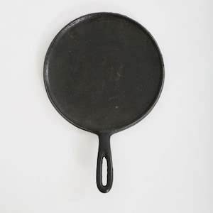 May include: A black cast iron griddle pan, featuring a circular cooking surface and a handle with a hanging hole. The pan is designed for stovetop use and has a rustic, well-used appearance. The pan is set against a white background.