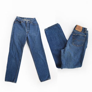 May include: Two pairs of blue denim jeans. One pair is laid flat, showing the front with button closure. The other pair is folded, revealing the back pockets and a brown leather patch. Both pairs have a classic, straight-leg design.
