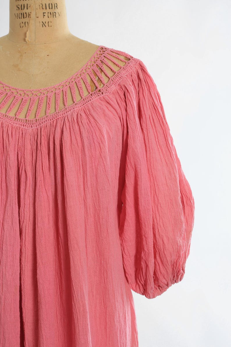 K&ouml;nnte beinhalten: Eine korallenfarbene Bluse mit H&auml;kel-Ausschnitt und langen, gerafften &Auml;rmeln. Der Stoff hat ein strukturiertes Aussehen, und der Ausschnitt weist ein detailliertes, offenes Design auf. Die Bluse ist einfarbig.