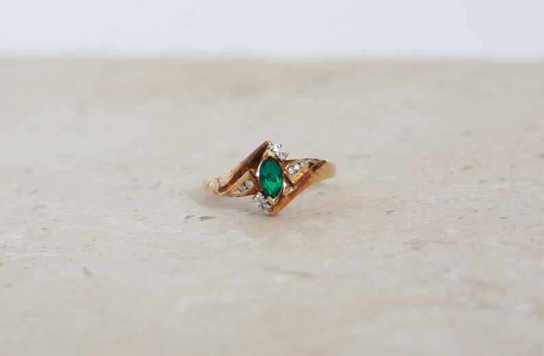 May include: A gold ring with a marquise-cut emerald center stone and small diamond accents. The ring has a unique, angular design. The emerald is a vibrant green, and the diamonds sparkle. A piece of jewelry.