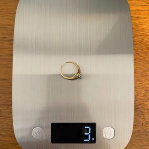 May include: A gold ring with a small turquoise stone is displayed on a digital kitchen scale. The scale reads 3 grams. The ring is centered on the scale, which has a brushed metal surface.