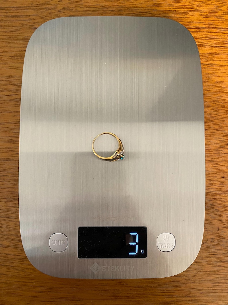 May include: A gold ring with a small turquoise stone is displayed on a digital kitchen scale. The scale reads 3 grams. The ring is centered on the scale, which has a brushed metal surface.