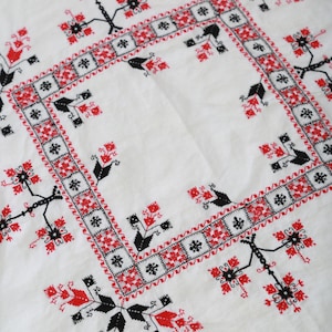 May include: White tablecloth with a red and black embroidered design. The design features a repeating pattern of stylized flowers and leaves.