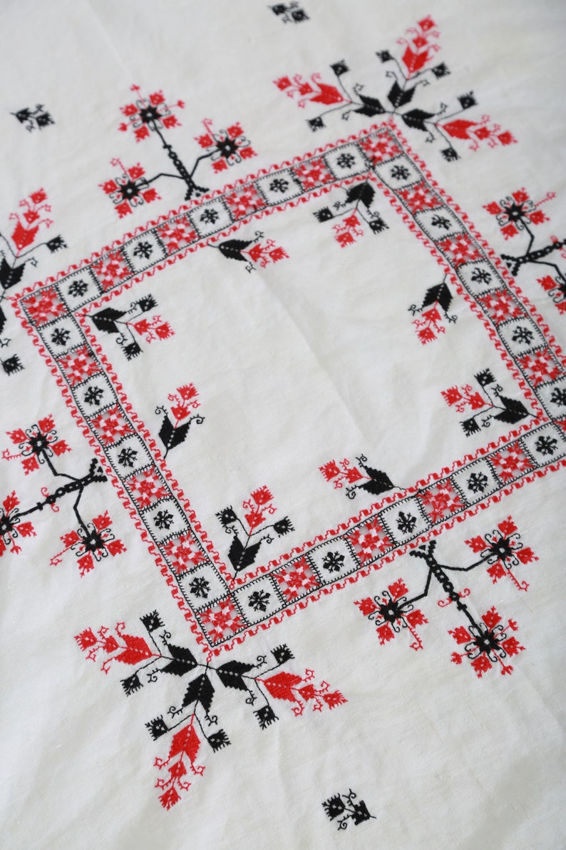 May include: White tablecloth with a red and black embroidered design. The design features a repeating pattern of stylized flowers and leaves.