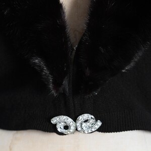 May include: A black fur collar with a black knit sweater underneath. The sweater has a zipper closure and a decorative rhinestone brooch.