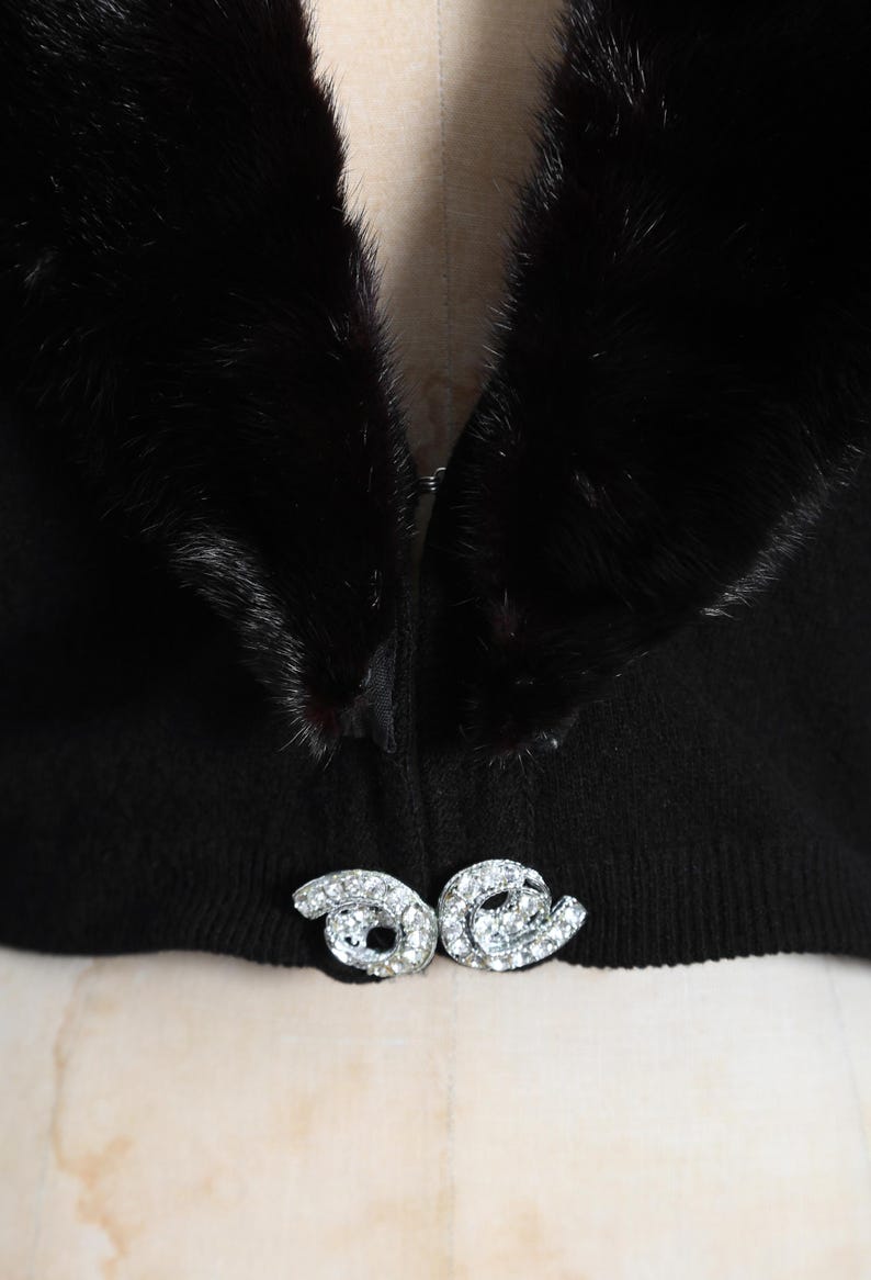 May include: A black fur collar with a black knit sweater underneath. The sweater has a zipper closure and a decorative rhinestone brooch.