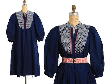 May include: A navy blue dress with white and red embroidered details. The dress has a round neckline, long sleeves, and a fitted waist with a red and white belt.
