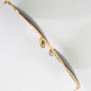 May include: A pair of sunglasses with thin gold-toned metal frames and dark, circular lenses. The eyewear features adjustable nose pads attached to the bridge and slender temples extending from the hinges, captured from a side-angle perspective.