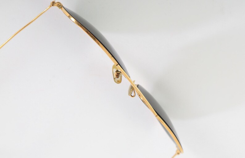 May include: A pair of sunglasses with thin gold-toned metal frames and dark, circular lenses. The eyewear features adjustable nose pads attached to the bridge and slender temples extending from the hinges, captured from a side-angle perspective.