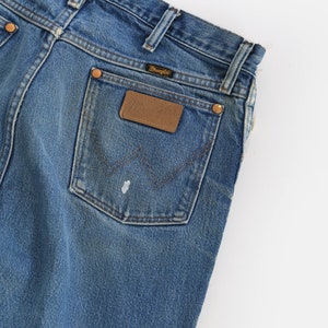 Op de afbeelding: Een paar blauwe denim jeans met een achterzak met een bruin lederen Wrangler-label.