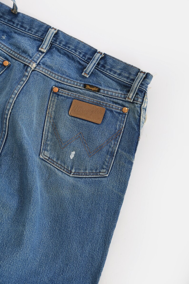 Op de afbeelding: Een paar blauwe denim jeans met een achterzak met een bruin lederen Wrangler-label.
