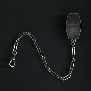 May include: A silver pocket watch chain with a rectangular fob. The chain is made of elongated links and has a clasp on one end. The fob has text on it. The background is black.