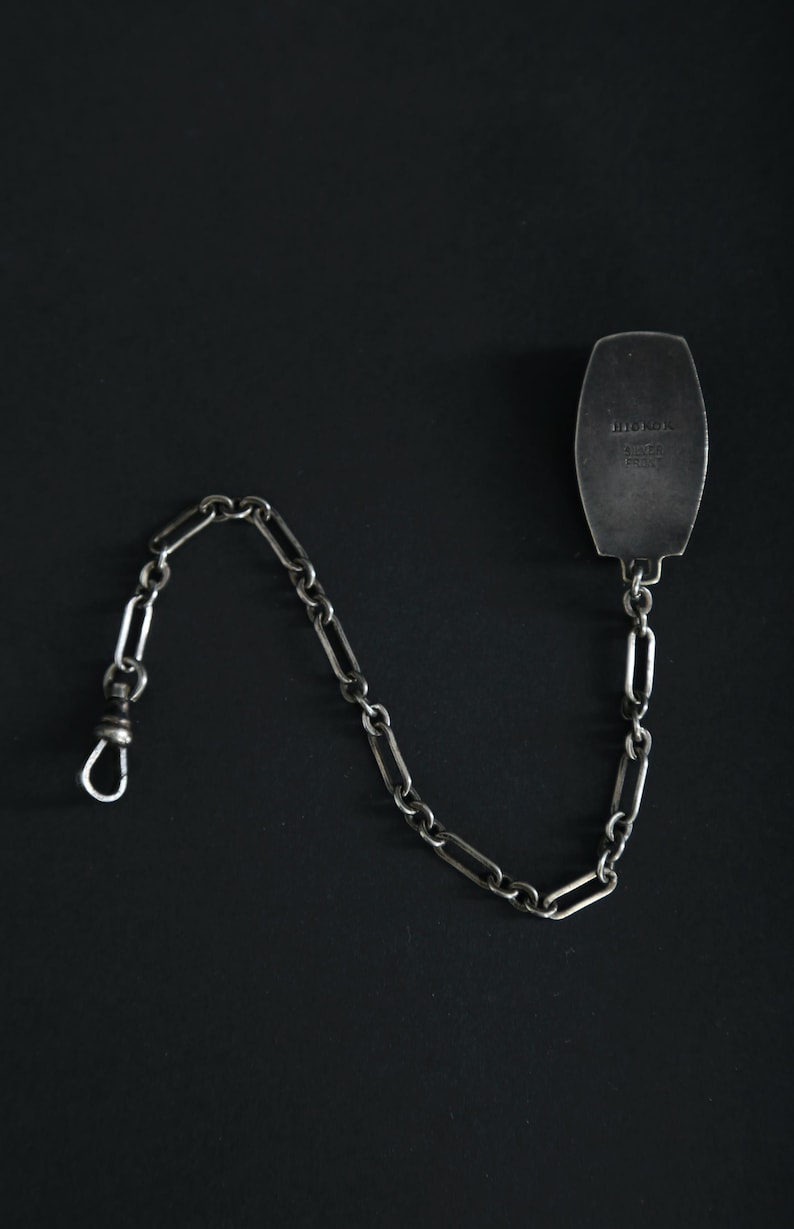 May include: A silver pocket watch chain with a rectangular fob. The chain is made of elongated links and has a clasp on one end. The fob has text on it. The background is black.