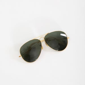May include: Aviator sunglasses featuring a thin gold-tone metal frame and dark green teardrop-shaped lenses. The design includes a double bridge across the top, adjustable nose pads, and slender temples, providing a classic look with a metallic finish.