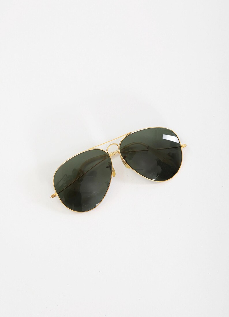 May include: Aviator sunglasses featuring a thin gold-tone metal frame and dark green teardrop-shaped lenses. The design includes a double bridge across the top, adjustable nose pads, and slender temples, providing a classic look with a metallic finish.