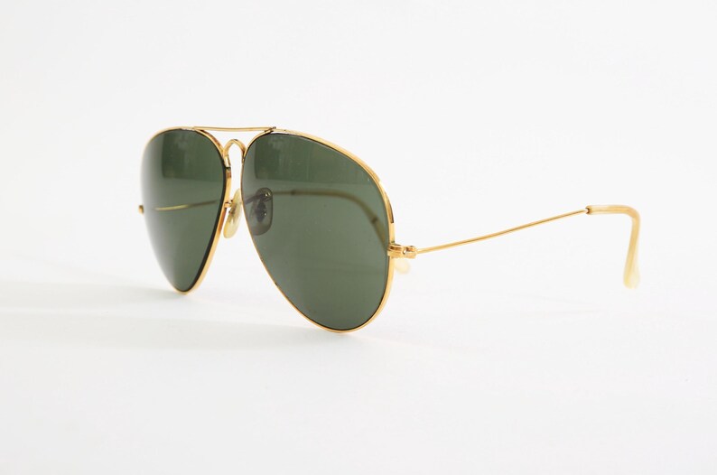 May include: Aviator sunglasses featuring a thin gold metal frame and dark green teardrop-shaped lenses. The design includes a double bridge across the top and slender gold temples with curved ends, providing a classic look with a metallic finish.