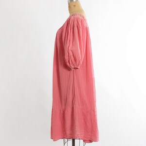 K&ouml;nnte beinhalten: Korallfarbenes Kleid mit quadratischem Ausschnitt und kurzen, gepufften &Auml;rmeln. Das Kleid hat eine lockere, flie&szlig;ende Silhouette und einen Stufenbund. Der Stoff scheint strukturiert zu sein, und das Kleid wird auf einer Schaufensterpuppe pr&auml;sentiert.