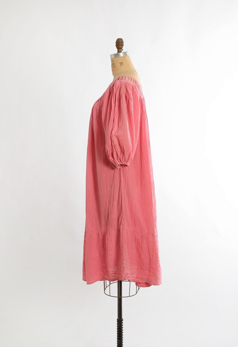 K&ouml;nnte beinhalten: Korallfarbenes Kleid mit quadratischem Ausschnitt und kurzen, gepufften &Auml;rmeln. Das Kleid hat eine lockere, flie&szlig;ende Silhouette und einen Stufenbund. Der Stoff scheint strukturiert zu sein, und das Kleid wird auf einer Schaufensterpuppe pr&auml;sentiert.