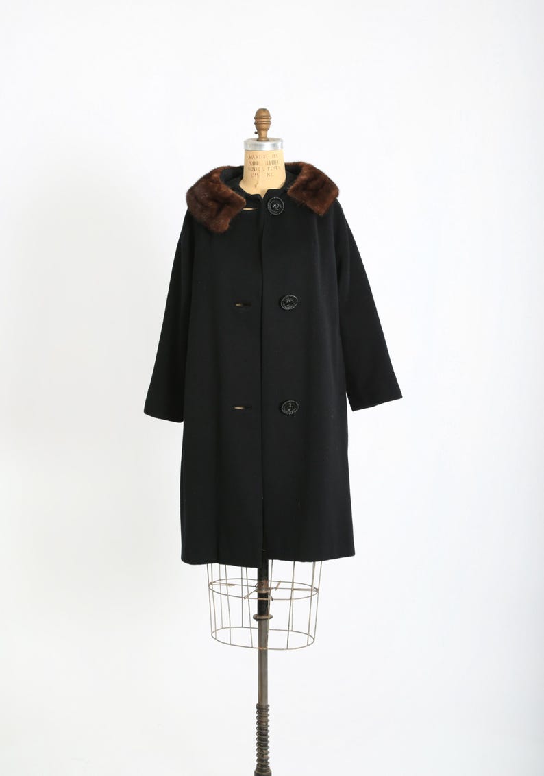 May include: A vintage black wool coat with a brown fur collar. The coat has a classic design with large buttons and a tailored fit. It is displayed on a dress form against a white background. The coat is a timeless piece of outerwear.