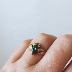 May include: A gold ring featuring a marquise-cut emerald-green gemstone as the centerpiece, flanked by smaller clear gemstones. The ring has a split shank design and is set against a neutral background.