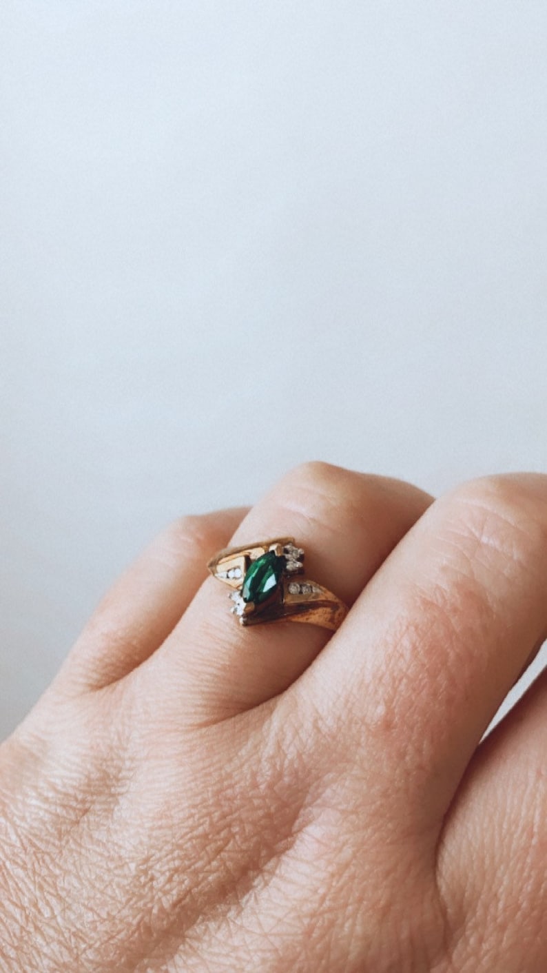 May include: A gold ring featuring a marquise-cut emerald-green gemstone as the centerpiece, flanked by smaller clear gemstones. The ring has a split shank design and is set against a neutral background.