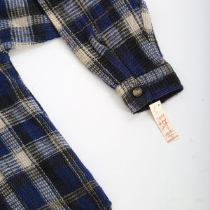 May include: Blue, black, and beige plaid flannel shirt with a button-up closure. The shirt has a waffle texture and a tag that reads "4897  Long Sleeve Shirt  12-14  $19.99".