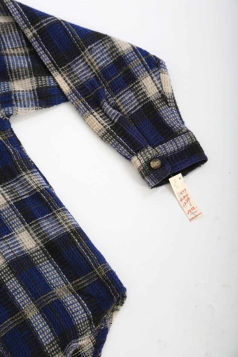 May include: Blue, black, and beige plaid flannel shirt with a button-up closure. The shirt has a waffle texture and a tag that reads "4897  Long Sleeve Shirt  12-14  $19.99".