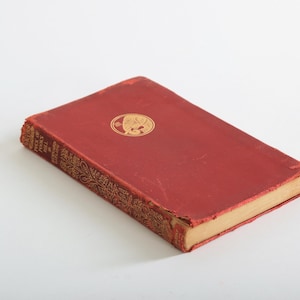 Puede incluir: Un libro antiguo de tapa dura roja con detalles dorados. Presenta un emblema dorado en la portada y patrones dorados decorativos en el lomo. Las páginas amarillentas sugieren antigüedad. Dimensiones aproximadas: 20 cm x 13 cm.