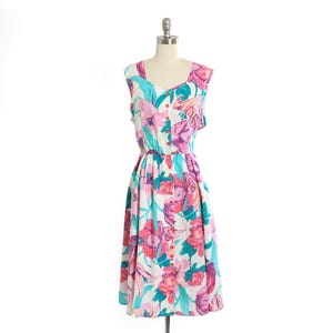May include: A sleeveless floral dress in white, pink, turquoise, and purple. The dress features a scoop neckline, button-up front, and a gathered waist. The dress is displayed on a mannequin.