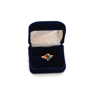 May include: A gold ring with an emerald-cut green gemstone and small white accent stones, displayed in an open, navy blue velvet ring box. The ring has a unique, angular design.