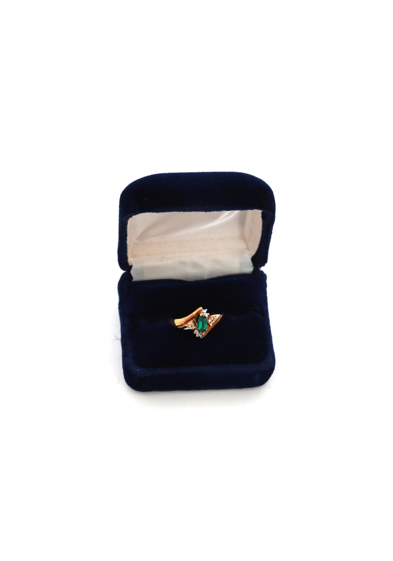 May include: A gold ring with an emerald-cut green gemstone and small white accent stones, displayed in an open, navy blue velvet ring box. The ring has a unique, angular design.