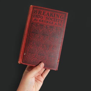 May include: A vintage book titled "Breaking Into Society" by George Ade. The book has a red cover with black decorative patterns and text. The book is held in a hand against a dark background.