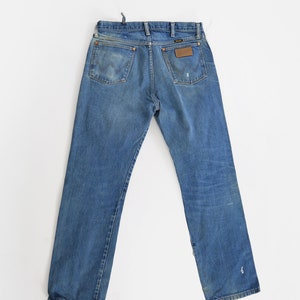 Op de afbeelding: Een paar blauwe denim jeans met een bruin leren patch op de achterzak met de tekst "Wrangler".