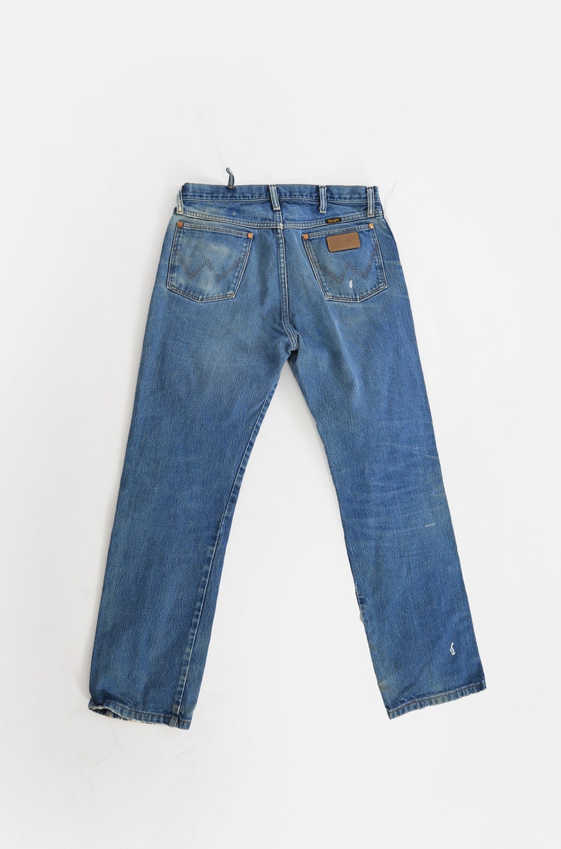Op de afbeelding: Een paar blauwe denim jeans met een bruin leren patch op de achterzak met de tekst "Wrangler".