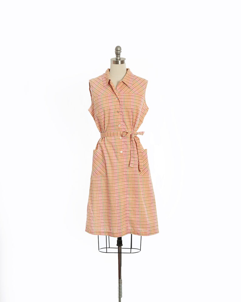 May include: A sleeveless dress with a plaid pattern in shades of pink, orange, and yellow. The dress has a collared neckline, button closure, and a belt.