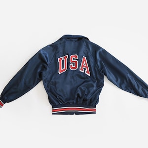 May include: A navy blue satin bomber jacket featuring the text USA in red letters with a white outline on the back. The jacket has a collar, long sleeves, and ribbed cuffs and a hem with red, white, and blue horizontal stripes.