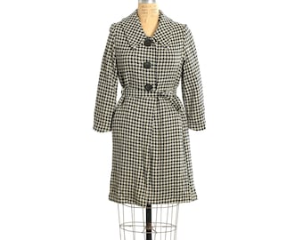 May include: A black and white houndstooth coat, shown on a wire dress form. The coat has a notched collar, three black buttons, and a matching belt. The coat has three-quarter sleeves and a knee-length hem. The coat is a vintage style.