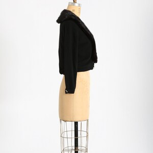 May include: A black cropped cardigan with a fur collar on a dress form. The cardigan has a shawl collar and is made of a soft, knit fabric.