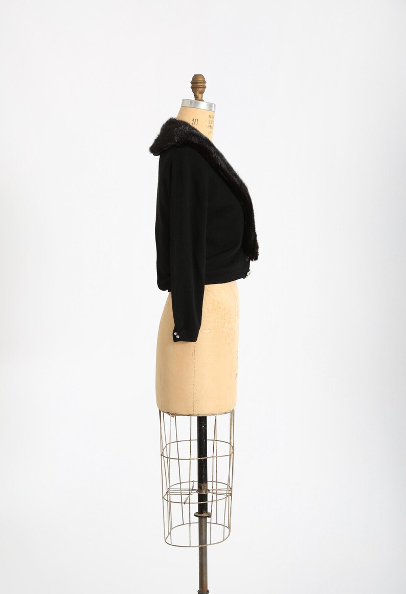 May include: A black cropped cardigan with a fur collar on a dress form. The cardigan has a shawl collar and is made of a soft, knit fabric.