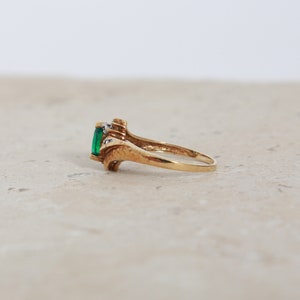May include: A gold ring featuring a rectangular emerald-cut green gemstone. The ring has a decorative design with small accent stones on either side of the emerald. The ring is shown on a neutral surface.