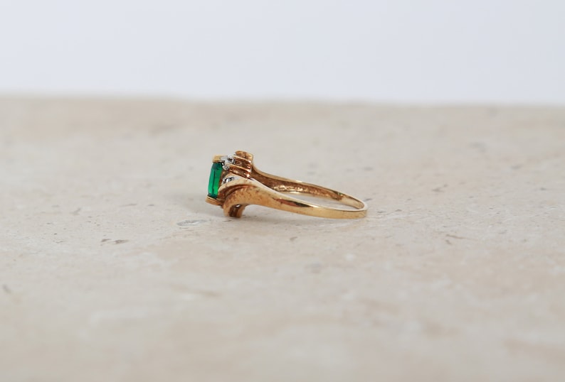 May include: A gold ring featuring a rectangular emerald-cut green gemstone. The ring has a decorative design with small accent stones on either side of the emerald. The ring is shown on a neutral surface.