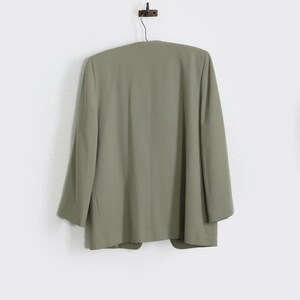 K&ouml;nnte beinhalten: Salbeigr&uuml;ner Blazer, der an einer Wand h&auml;ngt. Der Blazer hat ein klassisches Design mit langen &Auml;rmeln und einer taillierten Passform. Die Farbe ist ein ged&auml;mpfter, erdiger Ton, und das Kleidungsst&uuml;ck scheint aus einem glatten Stoff zu bestehen. Der Blazer h&auml;ngt an einem dunklen Metallhaken.